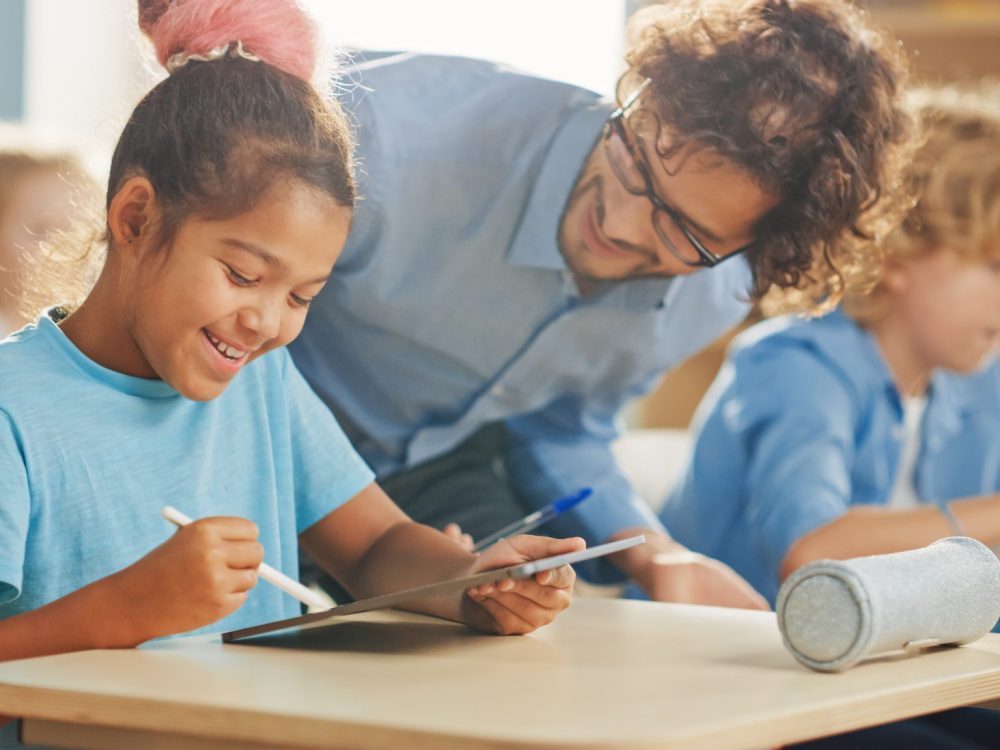 Elementary School Computer Science Class: Smart Girl Uses Digital Tablet Computer, Friendly Teacher Helps Her by Explaining Lesson and Asignment. Children Getting Modern Education